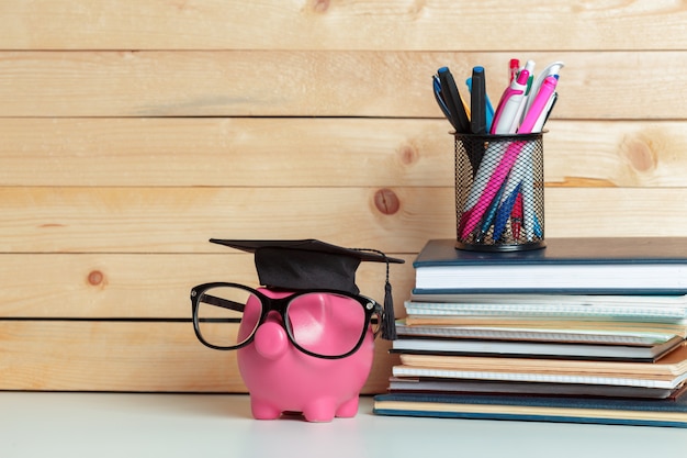 A stack of textbooks with a graduation cap on top, with a calculator and a pen next to the stack. In the background, there's a blurred image of a classroom setting.